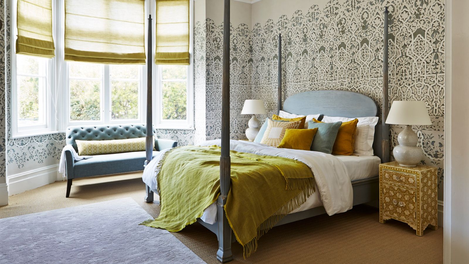 Four-poster bed in opulent room with patterned green wallpaper. The bed has neatly tucked in sheets, lots of throw pillows and a chesterfield sofa in the bay window
