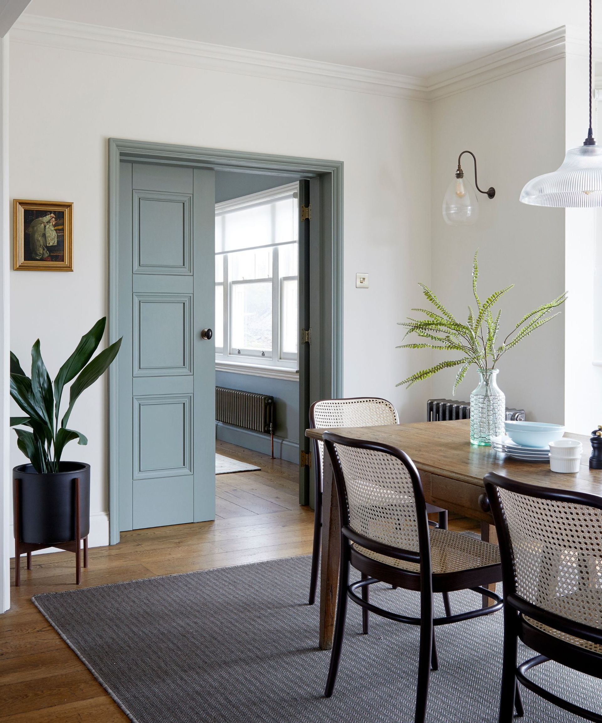 A cool toned dining room with panelled double doors leading to the next room. The doors are painted in an attractive light blue. There is a dining table with dining chairs and a grey rug on the wooden floorboards. There is a potted plant in the corner.