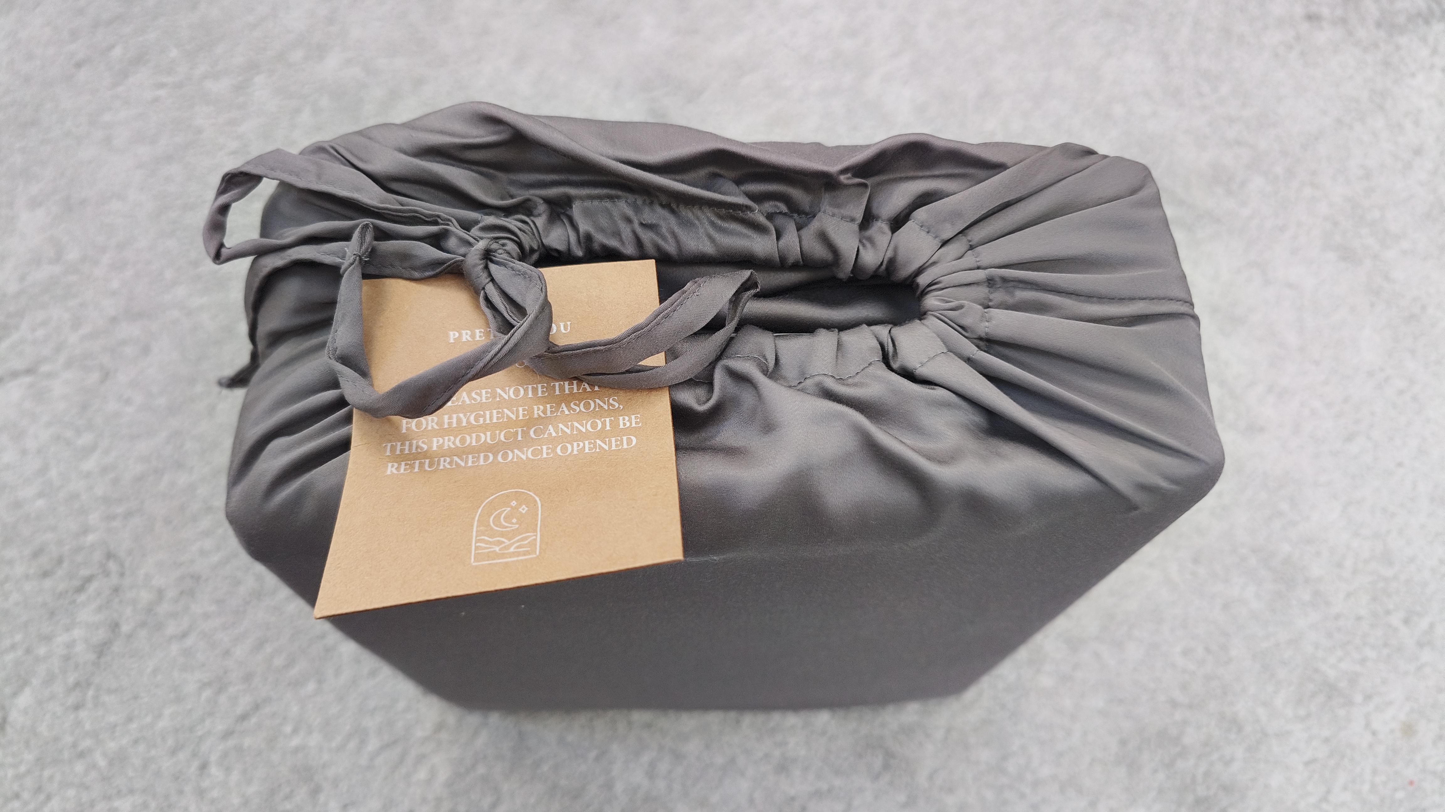 A charcoal-grey, silk-like bedding set is tightly packed inside a matching drawstring bag, featuring a brown card tag that notes return policies for hygiene reasons.