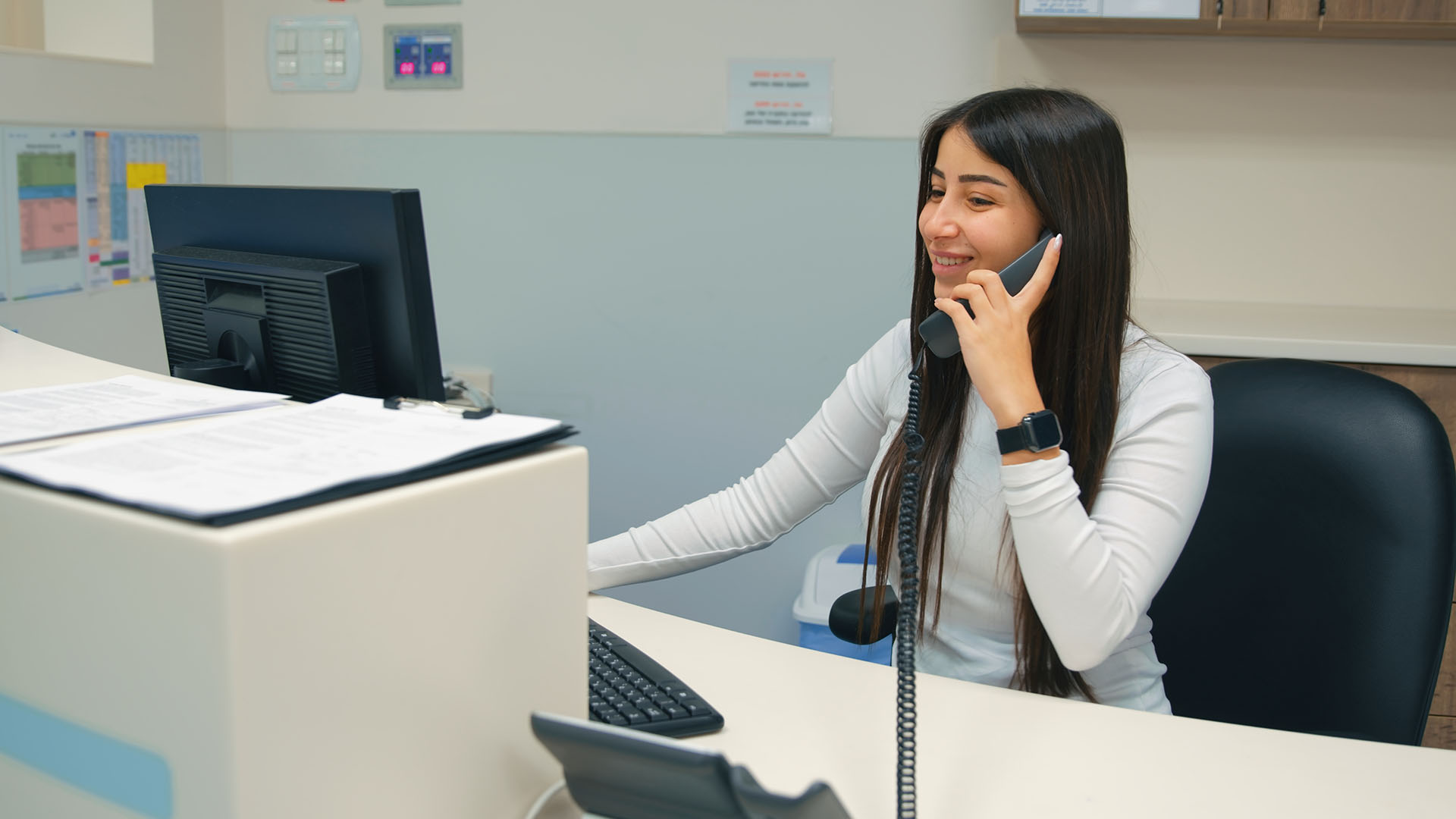 A receptionist taking a call on a landline while sitting at a desk and using a computer
