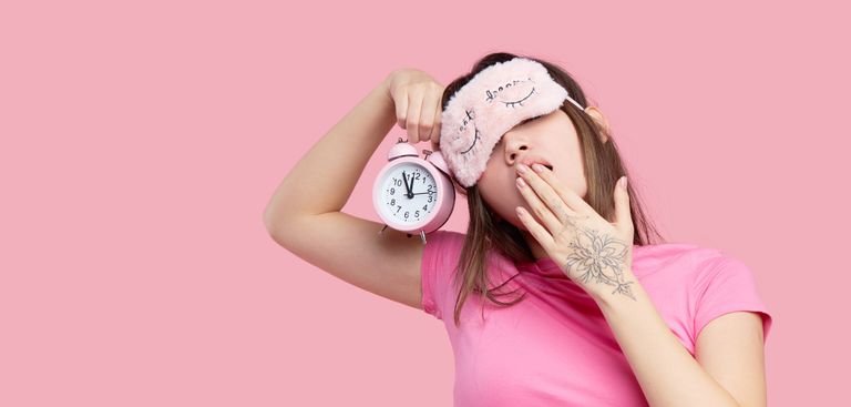 Close-up of yawning woman with sleep mask and alarm clock against pink background