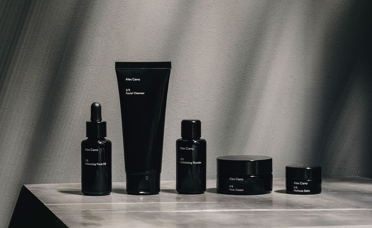 A display of Alex Carro&#039;s skincare capsule collection photographers on a vinyl surface against a grey wall. from left to right - A Black glass dropper bottle, a black cosmetic tube container, a black bottle with screw top and 2 round black pot jars in 2 different sizes