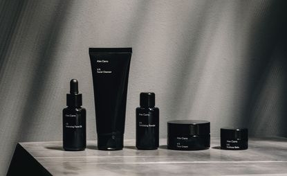 A display of Alex Carro's skincare capsule collection photographers on a vinyl surface against a grey wall. from left to right - A Black glass dropper bottle, a black cosmetic tube container, a black bottle with screw top and 2 round black pot jars in 2 different sizes