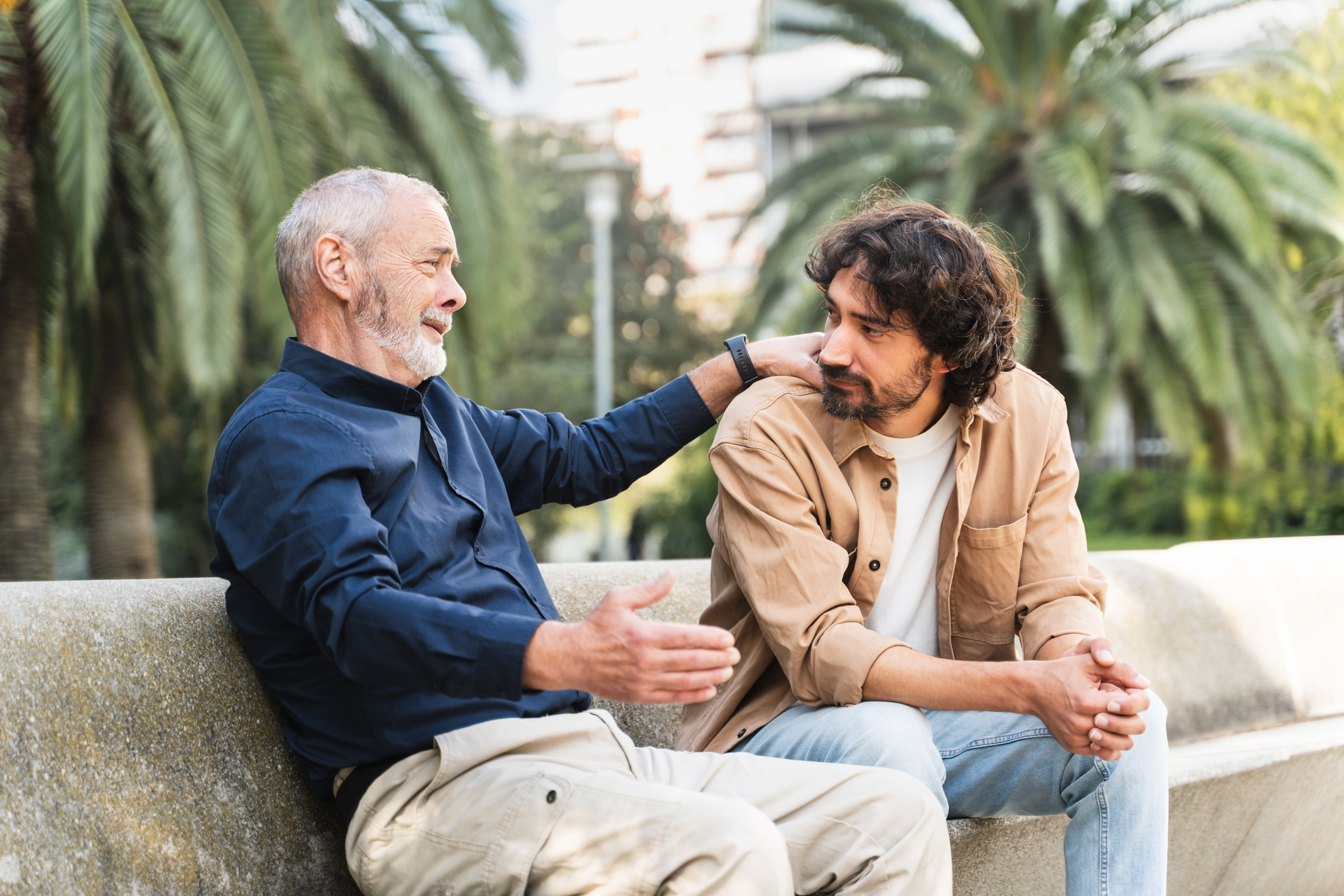 A father and his adult son sit outside on a bench talking.