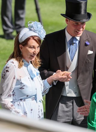 Carole Middleton wearing a pale blue floral dress and matching headband with a flower reaching holding Prince William's while standing in grass at Royal Ascot