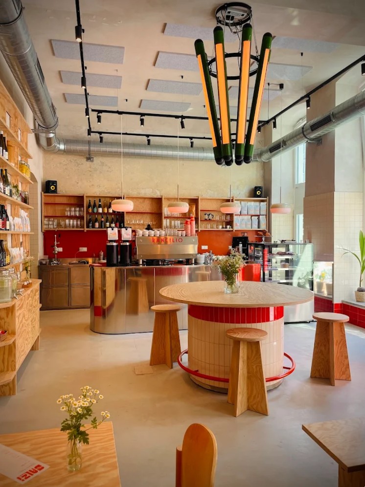 An industrial-style cafe features pale wood, geometrically cut furniture decorated with red accents in ceramic, pendant light with an oblong shape, a sculptural central chandelier with a green and orange structure, and visible air conducts in chrome.