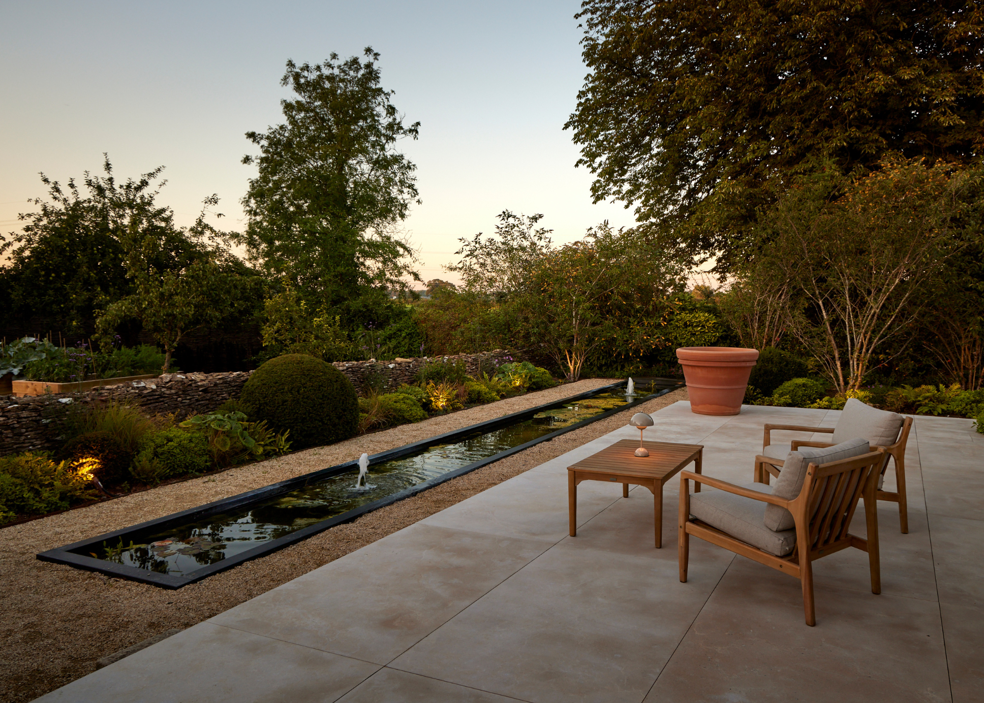 A long fountain outdoor water feature in a lush garden by a pair of armchairs and a wood side table