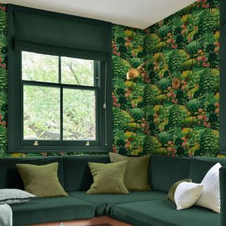 A living room with Arley Garden Day Wallpaper on a wall above a green velvet corner sofa. There's a window to the right, overlooking a tree.