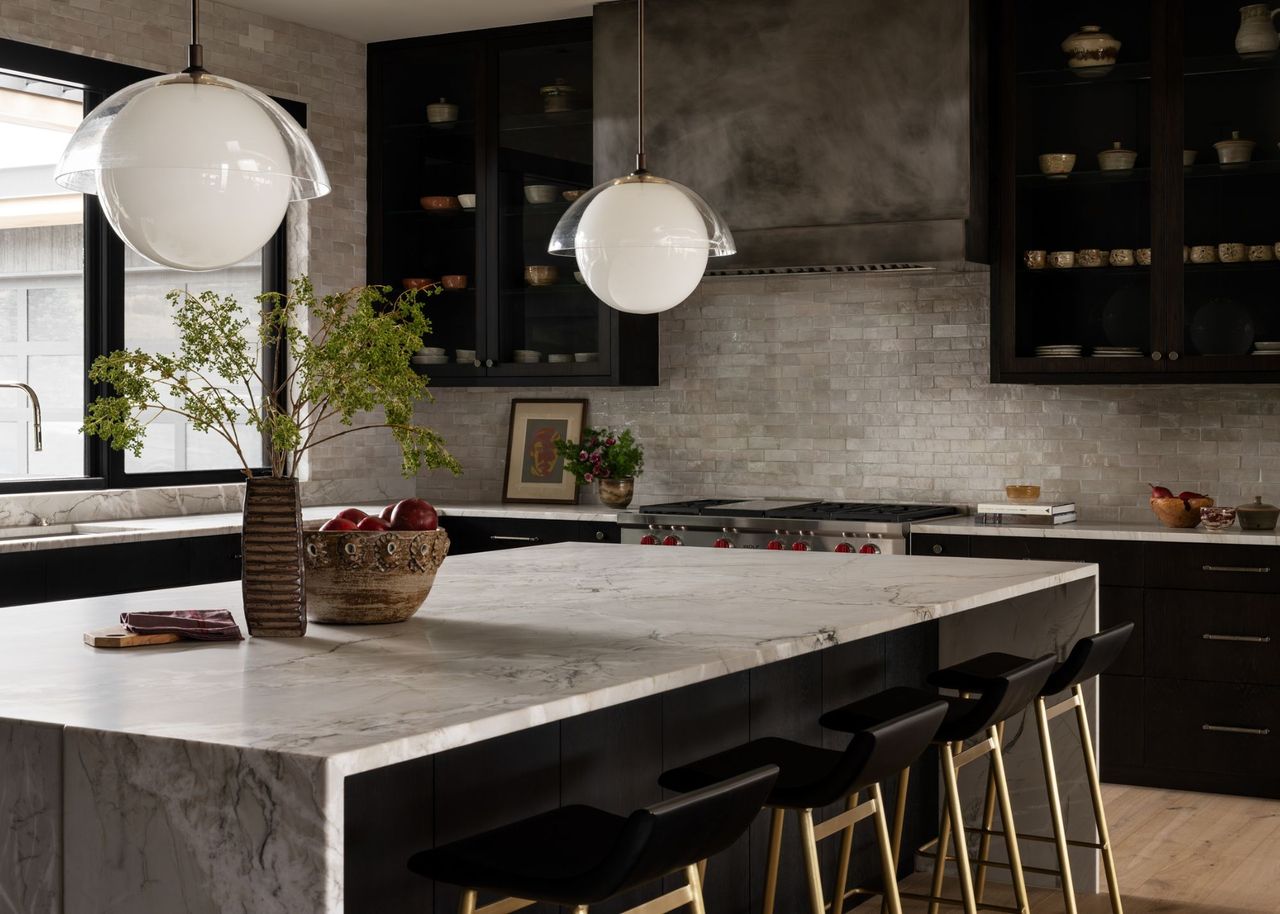 A contemporary kitchen with glass-fronted black cabinets, marble countertops and island and a gunmetal extractor hood with red knobs on the range cooker and round dome lights above the island