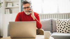 Mature man sitting on sofa at home and using smartphone and laptop for work.