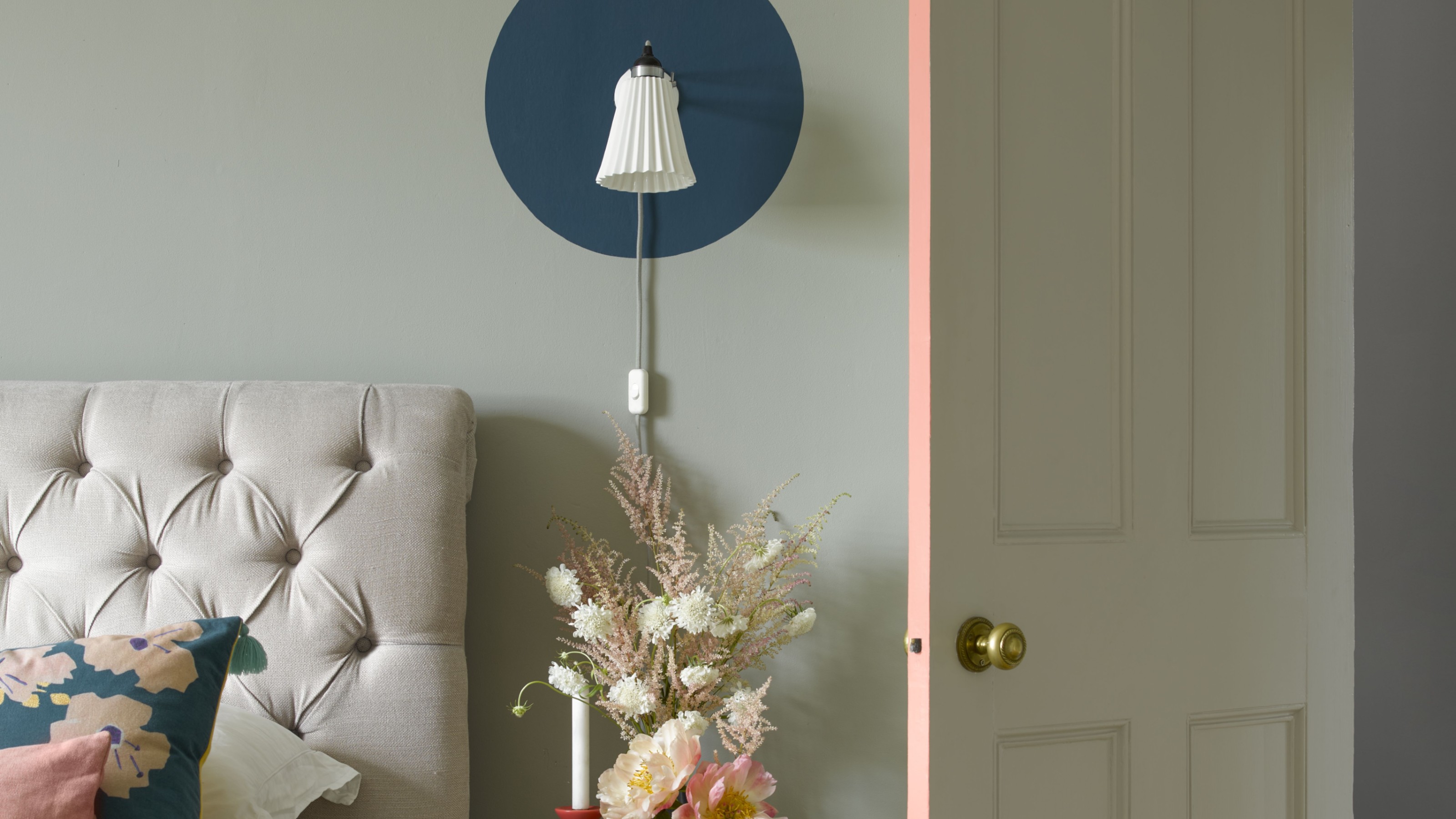 A soft green-painted bedroom with an upholstered headboard, a wall light highlighted by a navy blue circle painted around it and the edge of the door painted pink