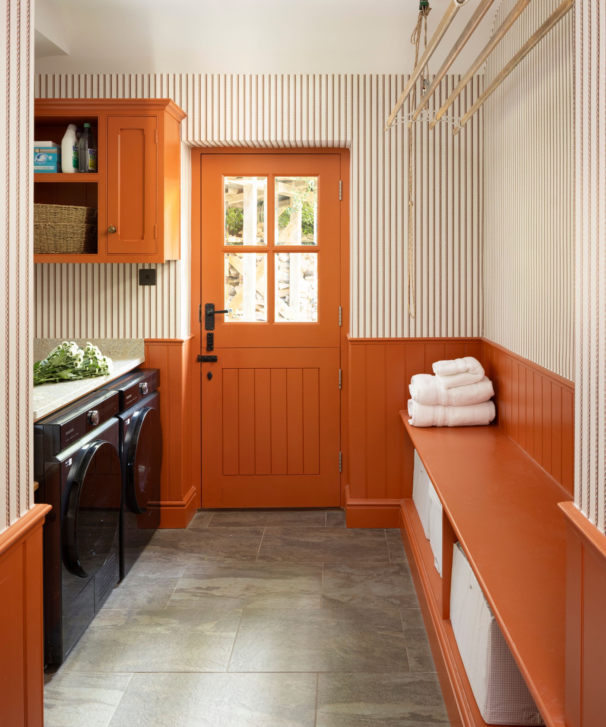 a bright orange laundry room with half paneled walls, painted door and trims and striped wallpaper with a mudroom style bench seat and washing machines