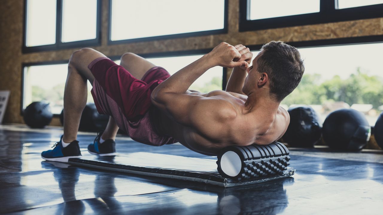 Young muscular Caucasian man massaging his upper back with a foam roller in an indoor gym