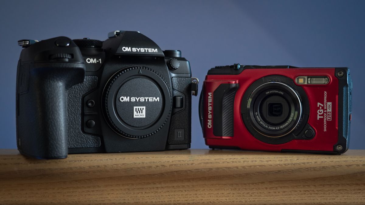 OM System OM-1 Mark II and OM System Tough TG-7 sat next to each other on a wooden surface, in front of a blue background