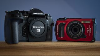 OM System OM-1 Mark II and OM System Tough TG-7 sat next to each other on a wooden surface, in front of a blue background