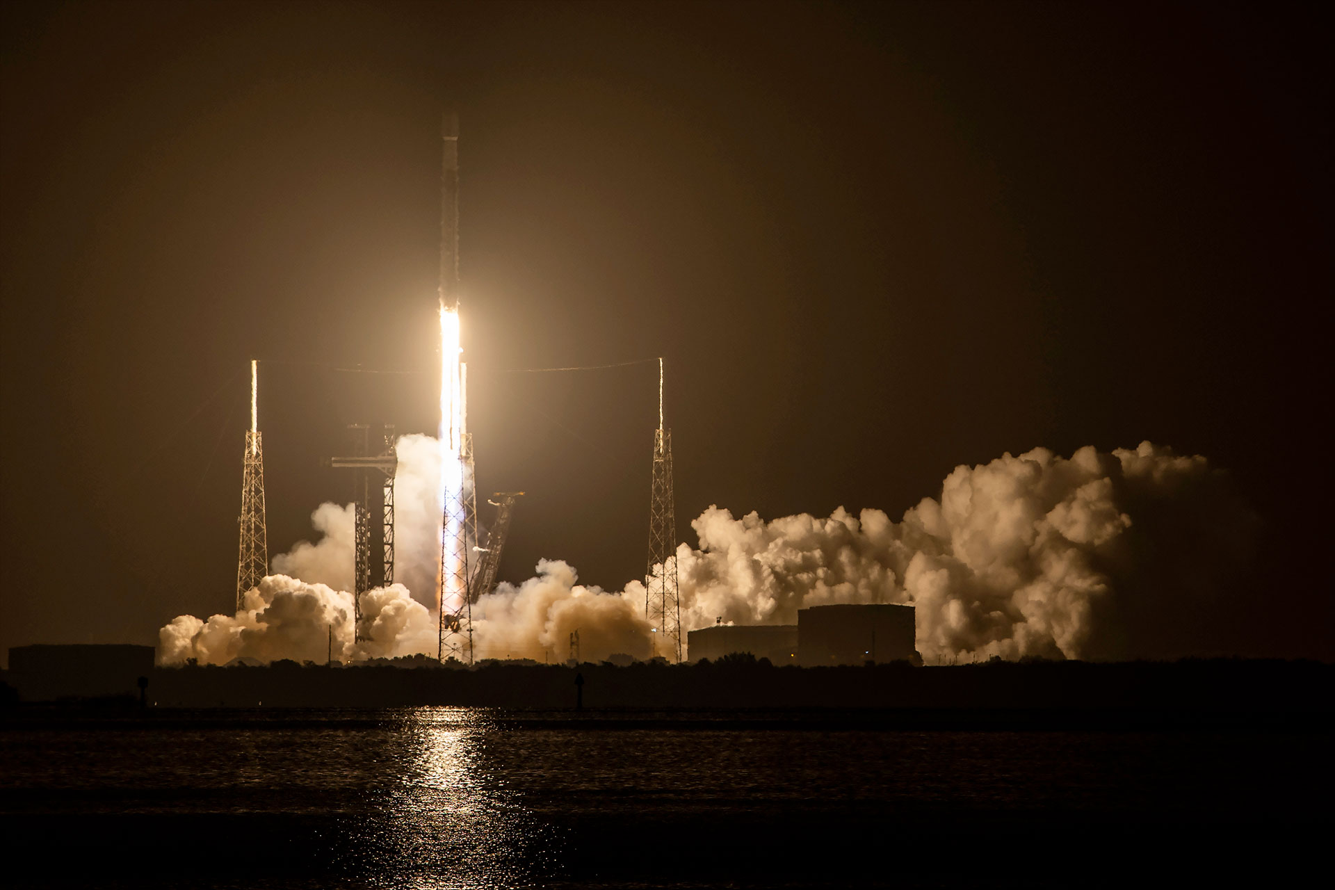 a rocket lifts off into the night sky, its orange-white thrust lighting up its launch pad below