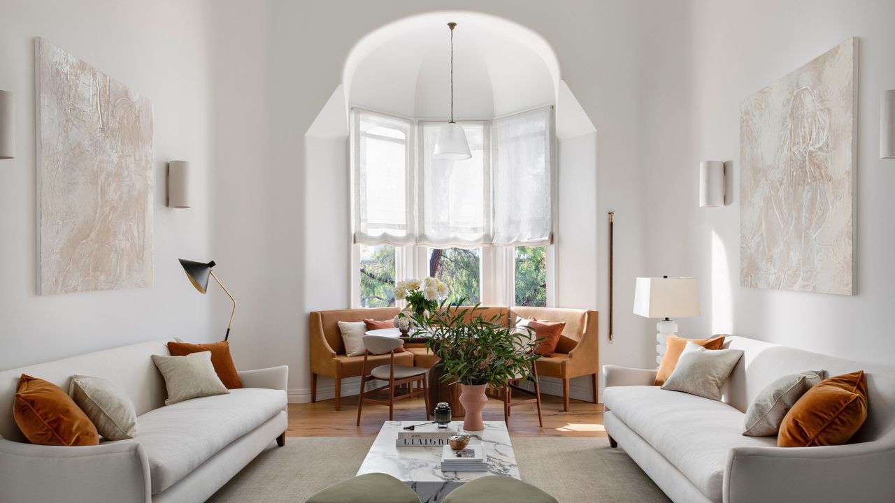 A large living room with oriel windows in a home. Next to the windows is a sofa shaped perfectly to fit by the window. On the right and left of the room are large sofas. 