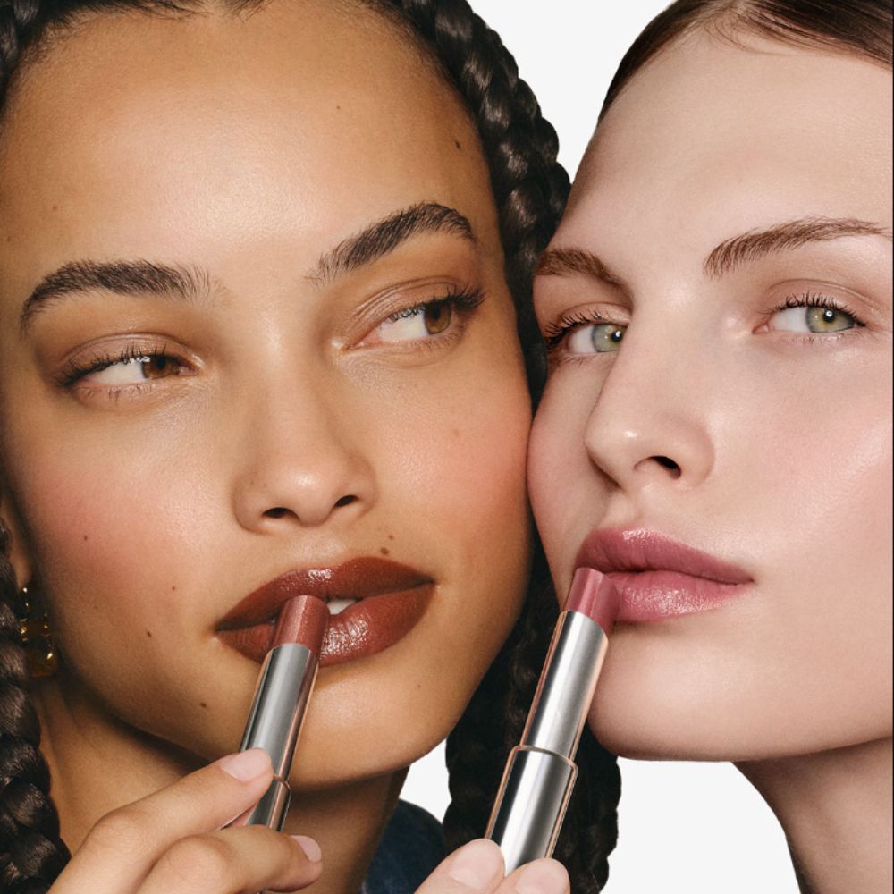 Two young woman applying Givenchy Beauty Rose Perfecto Shine Serum Stick, one looking at friend, the other at camera. 
