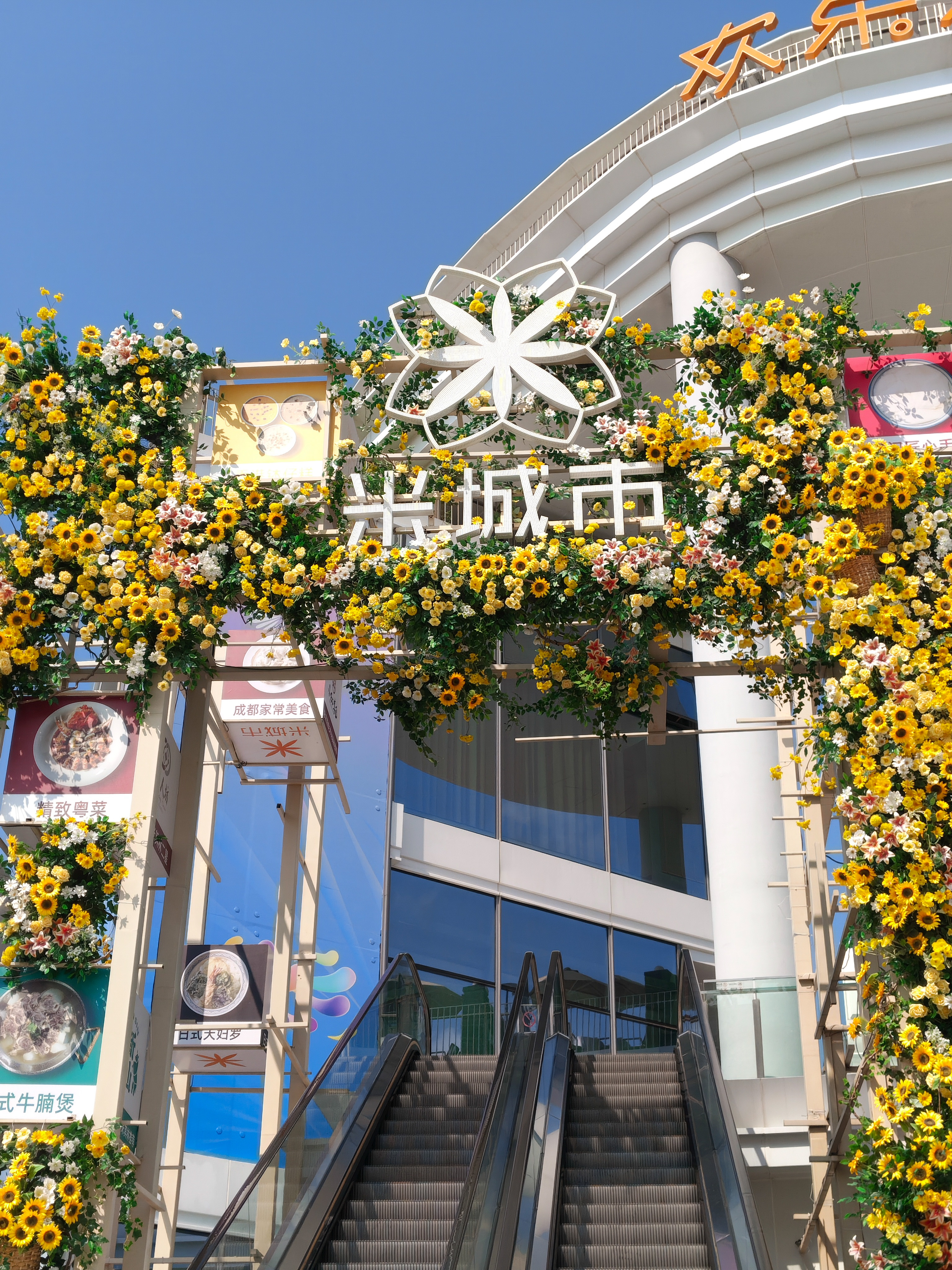 A wide shot of a modern building entrance featuring a pair of outdoor escalators leading upward. The entrance is decorated with a massive floral archway made of yellow and white flowers. At the top of the arch is a large, white geometric flower symbol and Chinese characters. To the left, vertical banners show pictures of various food dishes.