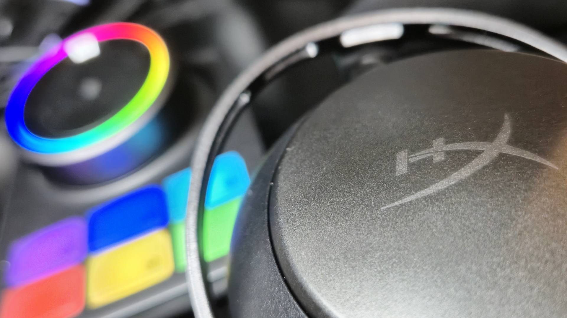 Close up of the side of a headset showing the HyperX logo, with a rainbow lit up base station in the background.