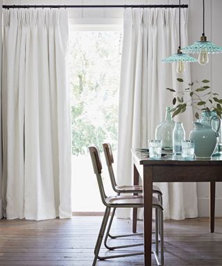 Sheer dining room curtains over floor to ceiling picture window with wooden dining table and chairs with decorative blue tableware and lights