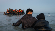 Two people standing in water, facing away from the camera, in front of a group of migrants in life vests on a small boat