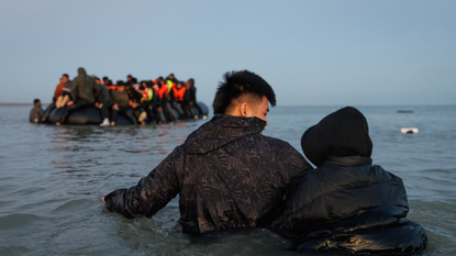 Two people standing in water, facing away from the camera, in front of a group of migrants in life vests on a small boat