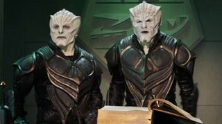 two people who appear to be white-skinned humanoid aliens with bony ridges on their faces stand in black armor