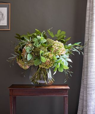 Bouquet of flowers in vase on wooden side table, dark green painted walls, gray curtain, hanging picture frame in corners