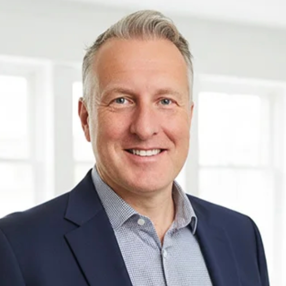 Smiling grey-haired man in a navy blazer and checked shirt, headshot in a bright indoor setting.