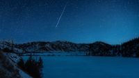 A shooting star is pictured streaking down through a starry sky above a frozen lake it night, bordered by snow-dappled hills.