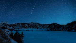 A shooting star is pictured streaking down through a starry sky above a frozen lake it night, bordered by snow-dappled hills.