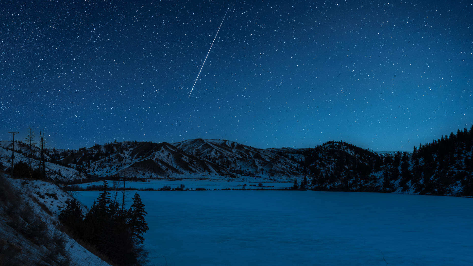 A shooting star is pictured streaking down through a starry sky above a frozen lake it night, bordered by snow-dappled hills.