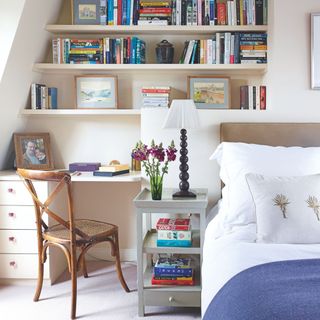 White painted bedroom with a bed, bedside table, and a desk with shelves full of books above it