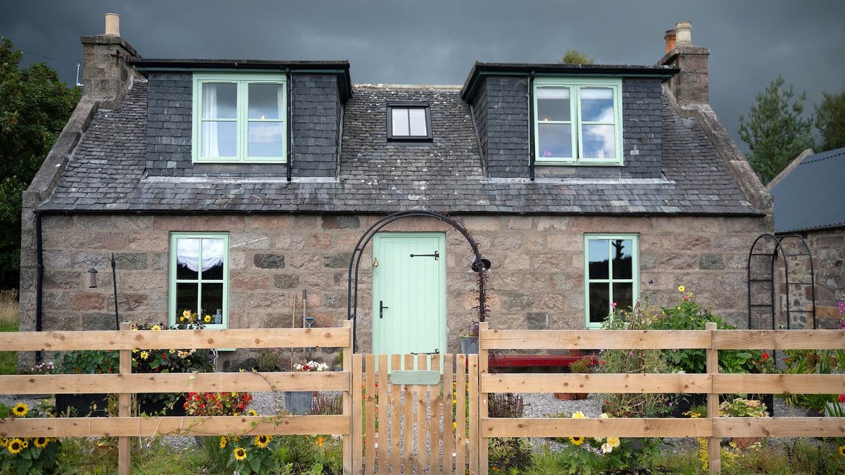 A stone wall cottage with dormer windows and a wooden fence in front of a small front garden