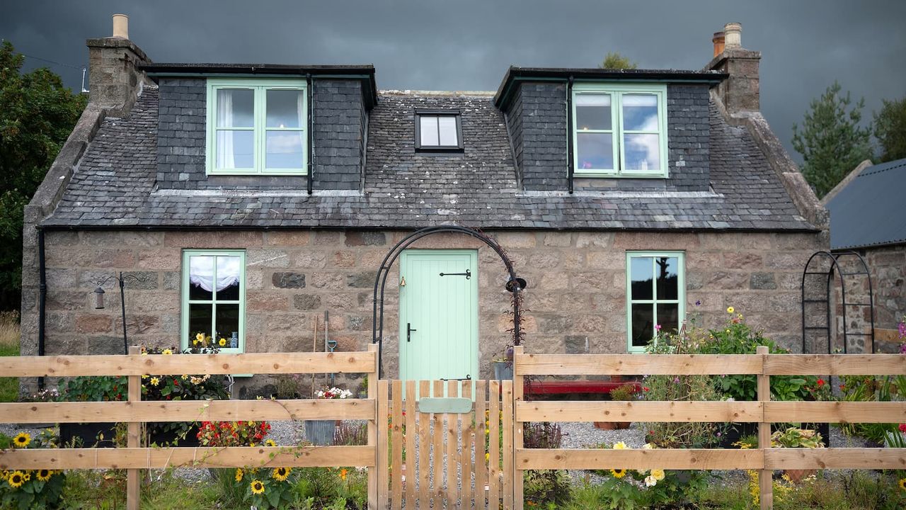 A stone wall cottage with dormer windows and a wooden fence in front