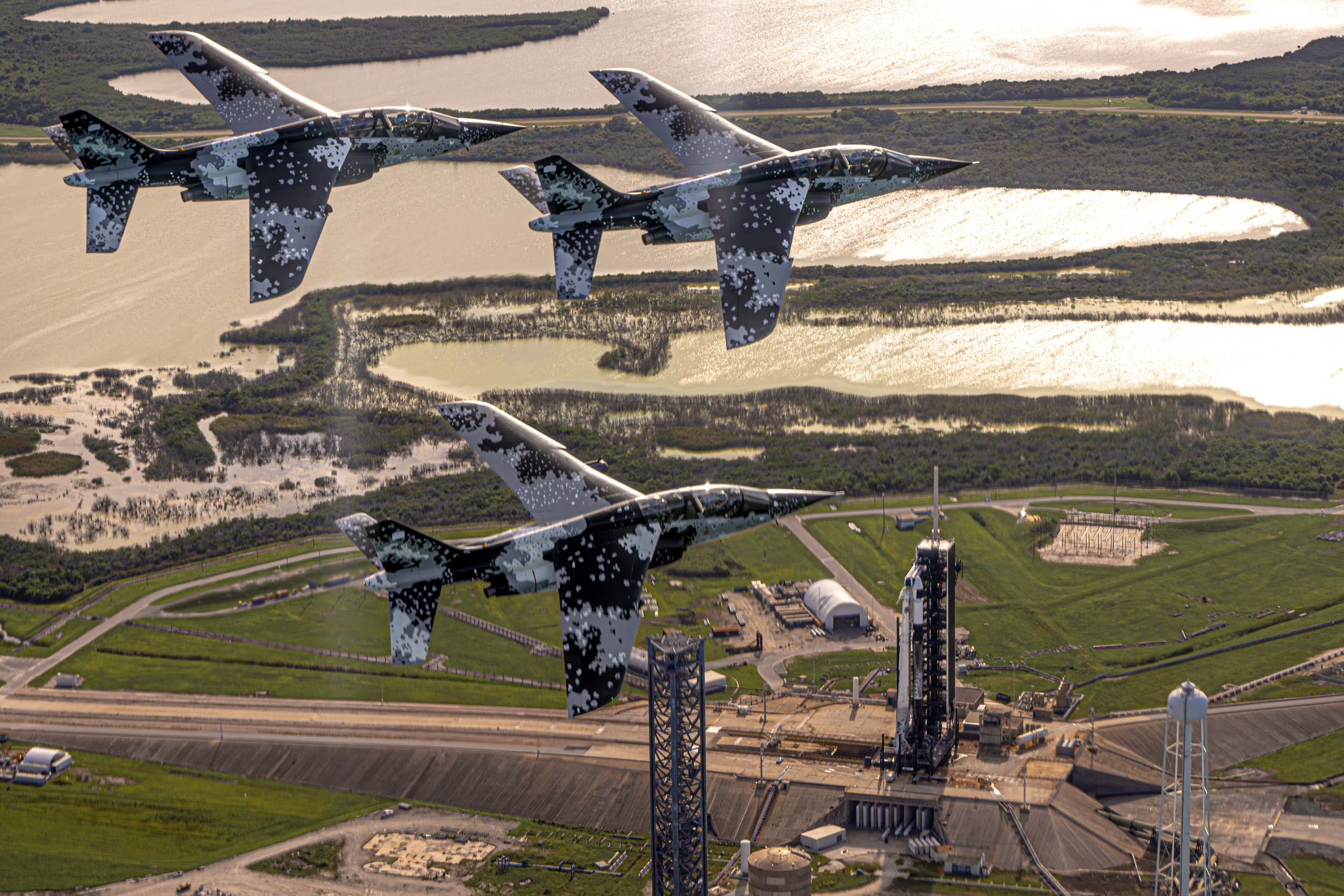 Three white, black, and grey camo jets fly over a rocket erect at a launchpad.