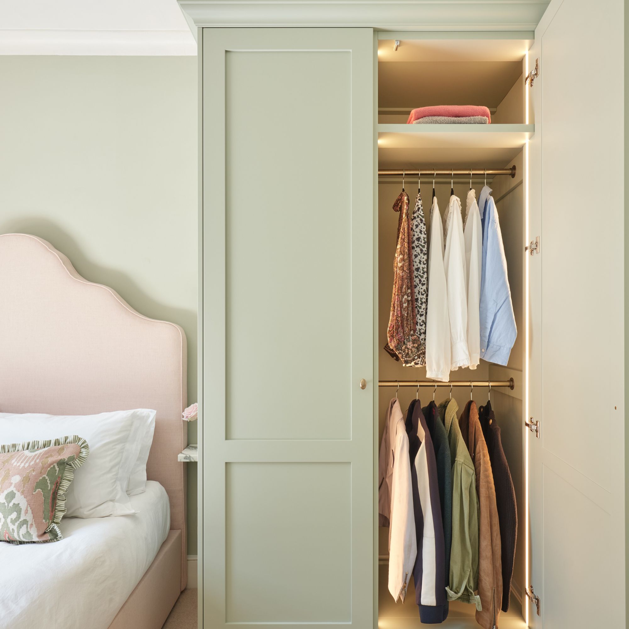 small green fitted wardrobe with one door open to reveal two hanging rails next to bed with pink headboard