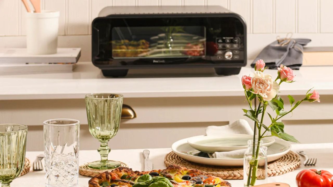 ProCook Electric Pizza Oven Air Fryer on the counter behind a table that&#039;s set with pizza and cutlery