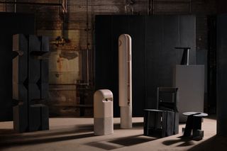 To the left, we see a black, high rectangle shape sculpture. In the middle, we see two beige sculptures with rounded tops. To the right, we see a black wooden chair with a wooden table next to it. It's all set in an industrial factory setting.