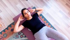 Woman performing crunch exercise at home. She lies on her back with her hands lightly touching the back of her head. She wears grey leggings and a black T-shirt