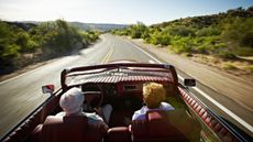 An older couple drive on a winding road in a convertible. 