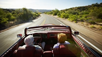 An older couple drive on a winding road in a convertible. 