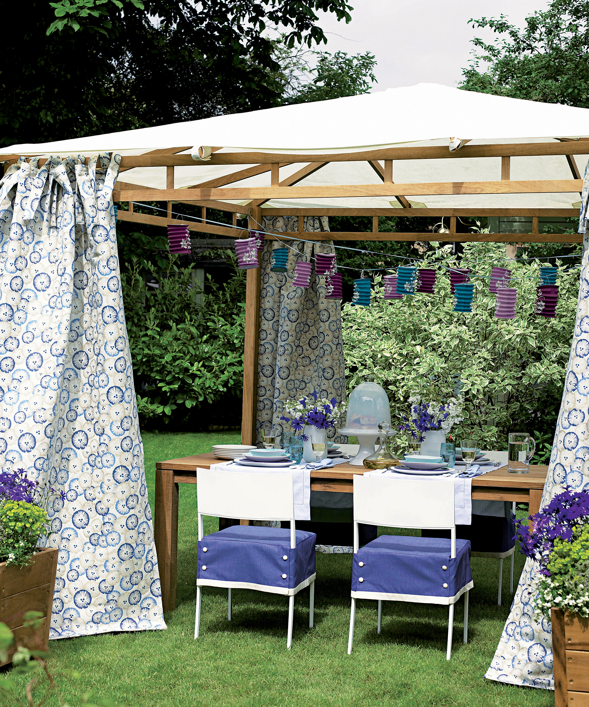 A gazebo in a garden with blue and white chairs and a table underneath