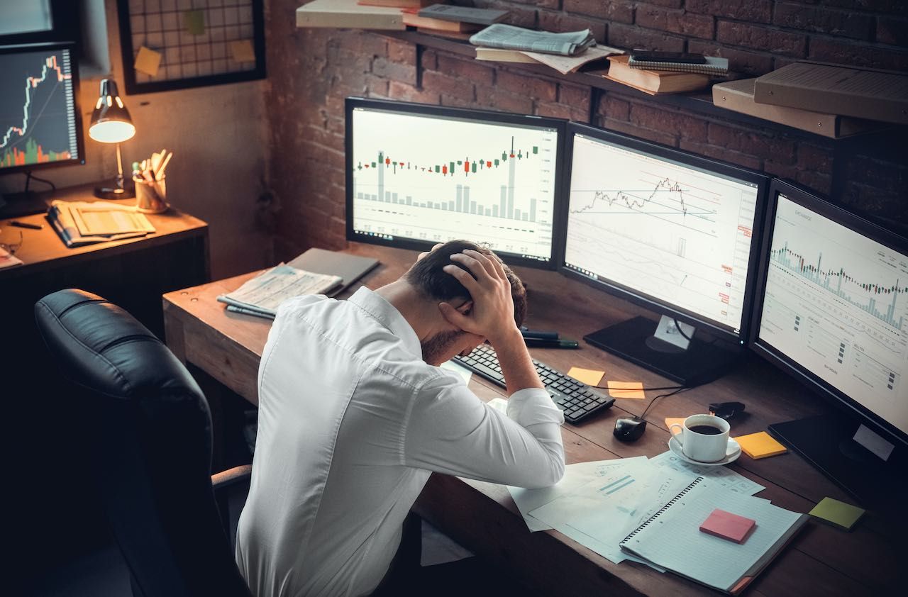 Young bearded man trader at office sitting at table holding head exhausted top view hard-working