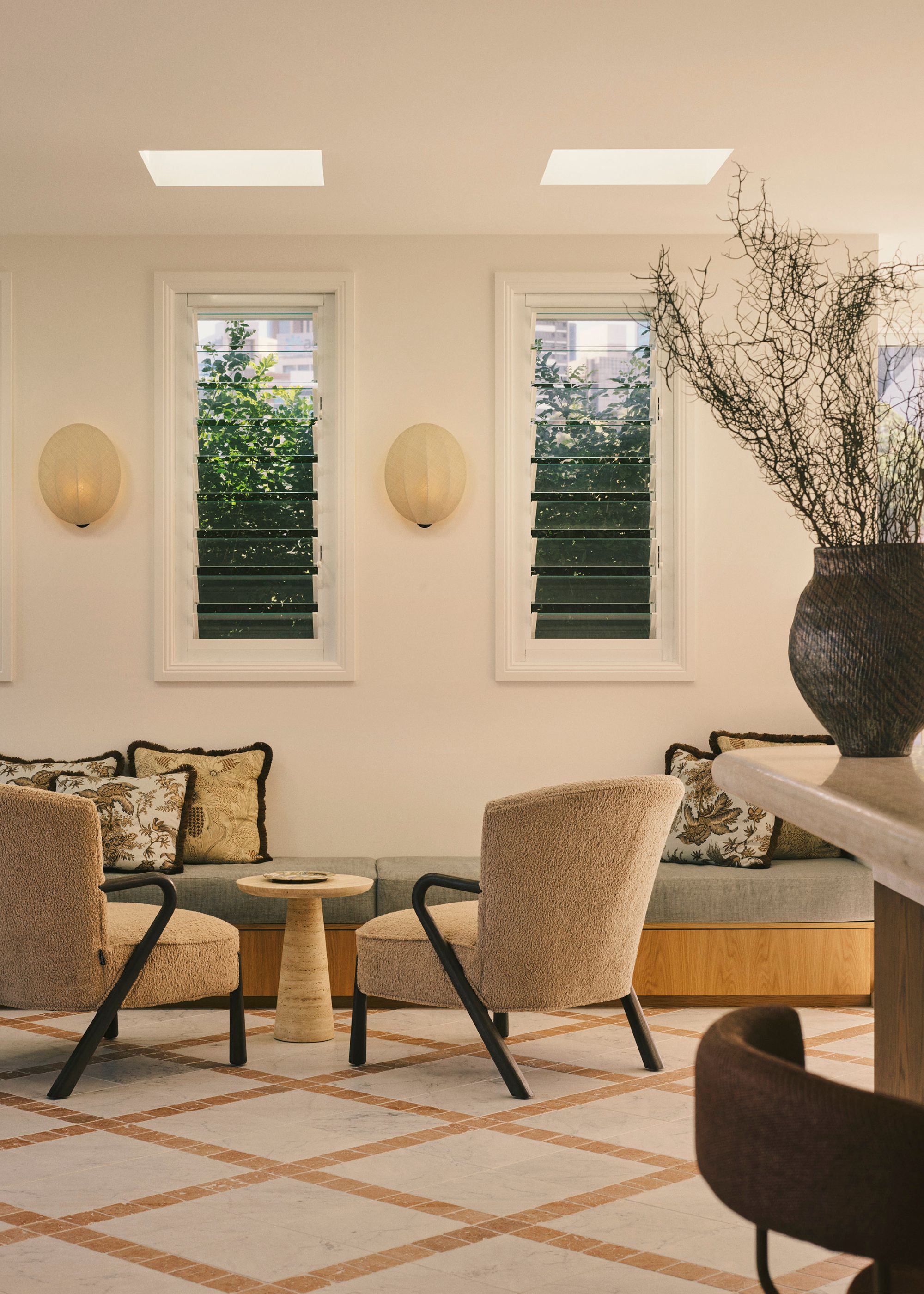 A white living room with beige armchairs, a blue bench, patterned cushions, a marble side table, a vase of stems, wall sconces, and skylights