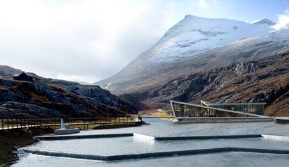 View of the Trollstigen Visitor Centre - a concrete and glass building with a geometric design surrounded by mountains and a river which is flowing over multiple levels
