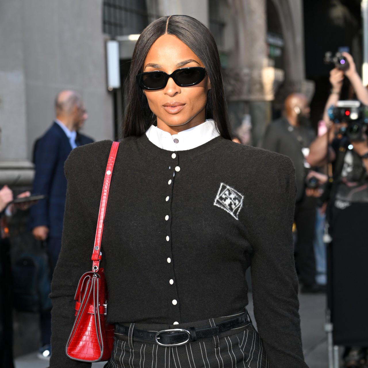 a photo of a black woman with long black hair wearing a black button-up cardigan with a white collar and a red purse. she is posing in front of a crowd of paparazzi