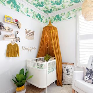White nursery with ceiling wallpapered in cactus print design, and an ochre canopy above the white cot