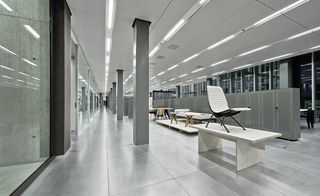 Open plan space in the G-Star RAW headquarters, Amsterdam, grey marble effect tiled floor, grey pillars, glazed walls, grey cabinets in a long row dividing the room, table and chairs, white coffee table with a white padded chair placed on top, white tiled ceiling with strobe lights